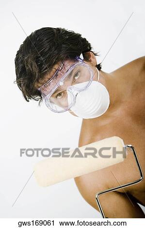 Stock Image - Portrait of a mid adult man wearing a pollution mask with a protective eyewear and holding a paint roller. Fotosearch