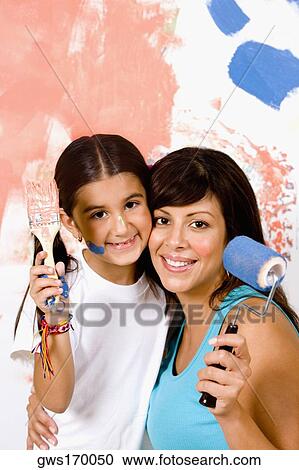 Stock Image - Portrait of a young woman holding a paint roller with her daughter holding a paintbrush. Fotosearch