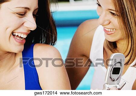 Close-up of a young woman holding a mobile phone and smiling with her friend View Large Photo Image Picture - Close-up of a young woman holding a mobile phone and smiling with her friend. Fotosearch