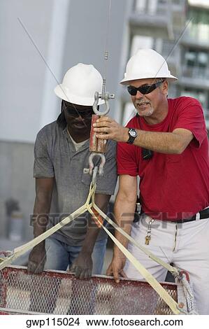 Picture - Two manual workers fastening a container in the hook of a lift. Fotosearch