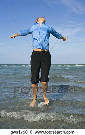 Stock Image - Mid adult woman jumping on the beach with her arms outstretched. Fotosearch
