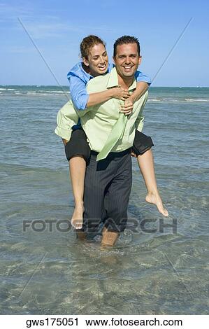 Stock Image - Mid adult woman riding piggyback on a mid adult man on the beach. Fotosearch