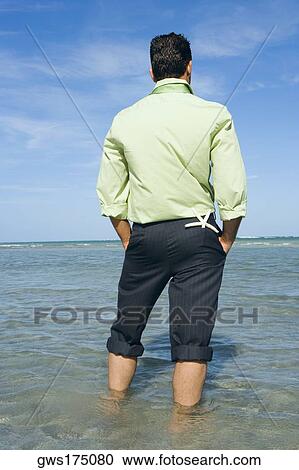 Stock Image - Rear view of a mid adult man with his hands in his pockets and standing on the beach. Fotosearch