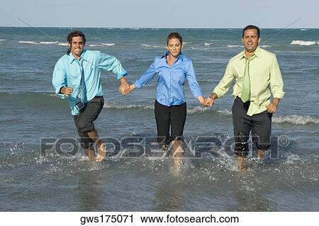 Stock Image - Two mid adult men running with a mid adult woman on the beach. Fotosearch