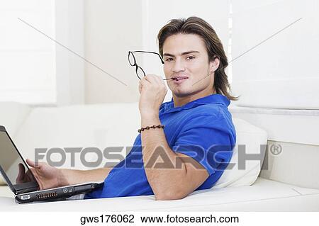 Stock Image - Portrait of a young man sitting in front of a laptop and holding eyeglasses. Fotosearch