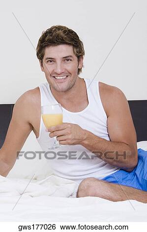 Stock Photograph - Portrait of a mid adult man sitting on the bed and holding a glass of orange juice. Fotosearch