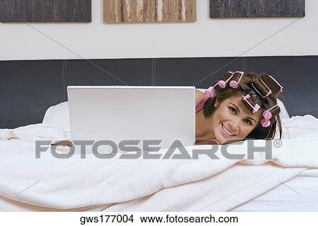 Picture - Portrait of a young woman wearing hair rollers and hiding herself behind a laptop on the bed. Fotosearch
