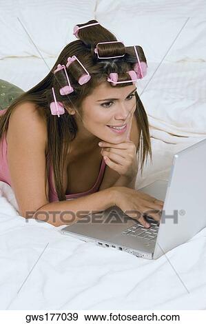 Stock Photo - Young woman wearing hair rollers and using a laptop on the bed. Fotosearch