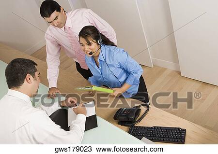 Stock Image - Rear view of a businessman writing with a businesswoman and a businessman looking at him. Fotosearch