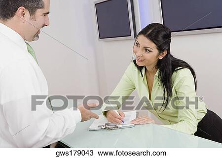 Receptionist writing on a clipboard with a businessman smiling in front of her View Large Photo Image Stock Image - Receptionist writing on a clipboard with a businessman smiling in front of her. Fotosearch