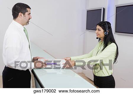 Stock Image - Side profile of a female customer service representative giving books to a businessman. Fotosearch