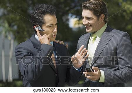 Stock Image - Businessman talking on a hands free device with another businessman standing beside him. Fotosearch