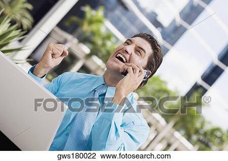 Businessman talking on a mobile phone and raising his fist in front of a laptop View Large Photo Image Stock Image - Businessman talking on a mobile phone and raising his fist in front of a laptop. Fotosearch