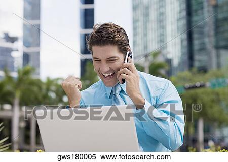 Stock Photography - Businessman talking on a mobile phone and raising his fist in front of a laptop. Fotosearch