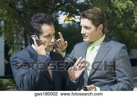 Stock Image - Close-up of a businessman talking on a hands free device with another businessman standing beside him. Fotosearch