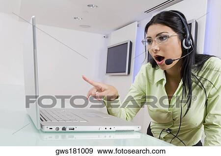 Stock Photo - Close-up of a young woman wearing a headset and pointing towards a laptop. Fotosearch