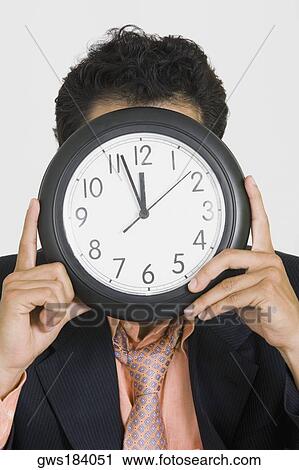 Stock Image - Close-up of a businessman hiding his face with a clock. Fotosearch