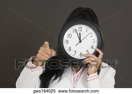 Stock Photography - Close-up of a businesswoman holding a clock in front of her face and pointing. Fotosearch