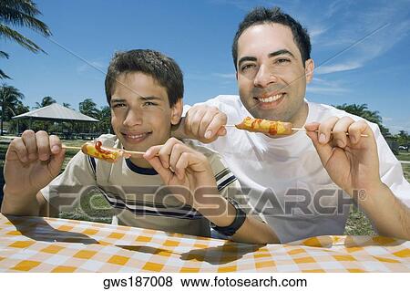 Stock Photo - Portrait of a mid adult man with his son eating sausages in a picnic. Fotosearch