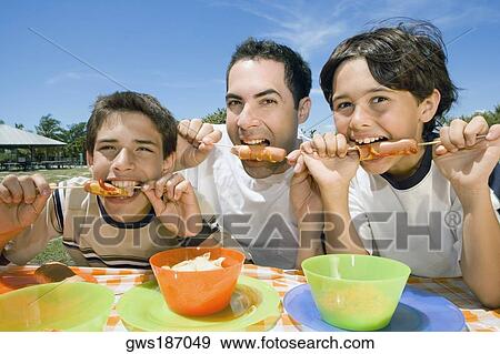 Portrait of a mid adult man with his two sons eating sausages in a picnic View Large Photo Image Stock Photo - Portrait of a mid adult man with his two sons eating sausages in a picnic. Fotosearch
