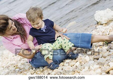Picture - Mid adult woman sitting on stones with her son at the water's edge. Fotosearch