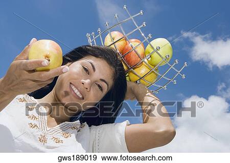 Stock Photo - Low angle view of a mid adult woman holding a fruit tray on her head and showing an apple. Fotosearch