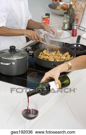 Stock Photograph - Close-up of a woman's hand pouring red wine into a wine glass with a maid preparing food in the background. Fotosearch