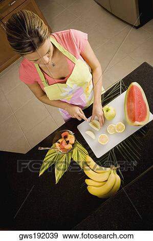 Stock Photo - High angle view of a young woman standing at the kitchen counter cutting fruits. Fotosearch