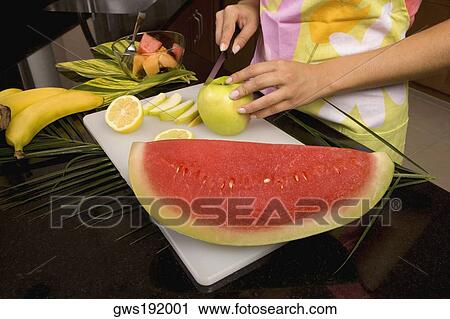 Stock Image - Mid section view of a woman standing at the kitchen counter cutting slices of an apple. Fotosearch