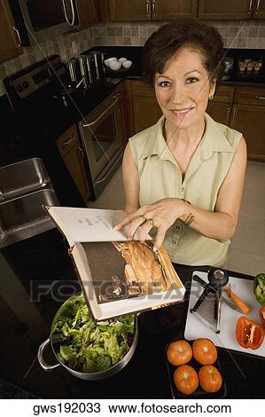 Stock Image - Portrait of a senior woman holding a cookbook in the kitchen and smiling. Fotosearch