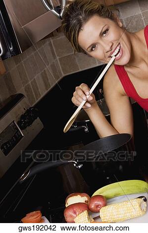 Stock Image - Portrait of a young woman holding a wooden spoon in her mouth and smiling in the kitchen. Fotosearch