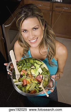 Stock Photography - Portrait of a young woman holding a wok of salad in the kitchen. Fotosearch