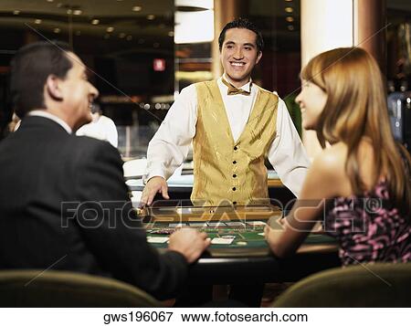 Stock Photo - Rear view of a mature man and a young woman sitting at a gambling table with a casino worker smiling in front of them. Fotosearch