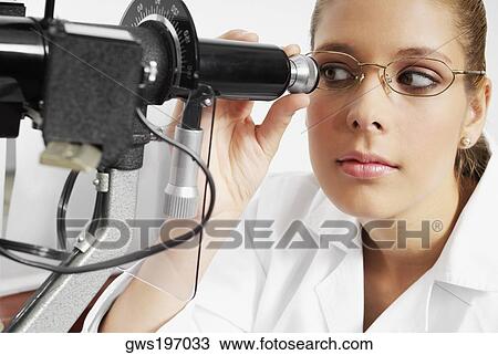 Stock Image - Close-up of a female optometrist looking through an eye test equipment. Fotosearch