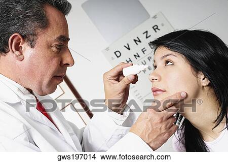 Picture - Side profile of a male doctor putting eye drops in a young woman's eye. Fotosearch