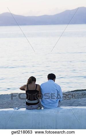 Stock Image - Rear view of a couple sitting on a wall near a beach, Mykonos, Cyclades Islands, Greece. Fotosearch