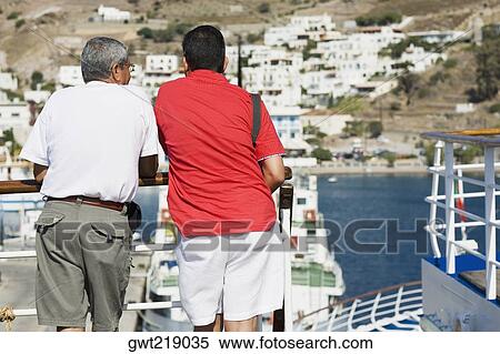 Stock Photography - Rear view of two men leaning against a railing of a yacht and looking at view, Patmos, Dodecanese Islands, Greece. Fotosearch