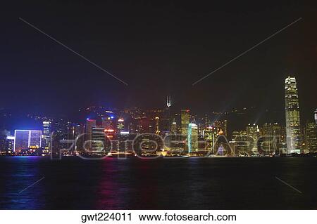 Stock Image - Skyscrapers lit up at night in a city, Victoria Harbor, Hong Kong Island, Hong Kong, China. Fotosearch