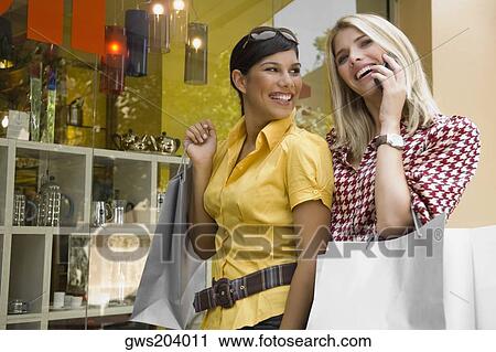 Young woman talking on a mobile phone with another young woman holding a shopping bag beside her View Large Photo Image Stock Image - Young woman talking on a mobile phone with another young woman holding a shopping bag beside her. Fotosearch