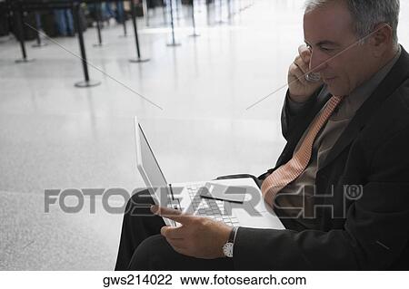 Stock Image - Side profile of a businessman talking on a mobile phone and using a laptop at an airport. Fotosearch
