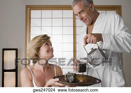 Senior man pouring tea from a tea kettle into the cup with a mature woman holding a tray View Large Photo Image Picture - Senior man pouring tea from a tea kettle into the cup with a mature woman holding a tray. Fotosearch