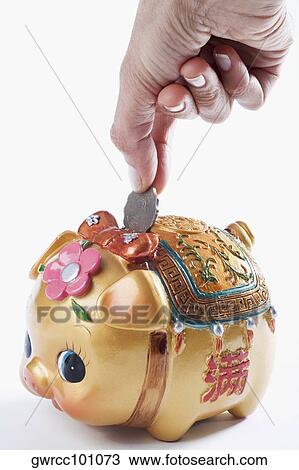 Stock Image - Close-up of a person's hand putting coin into a piggy bank. Fotosearch