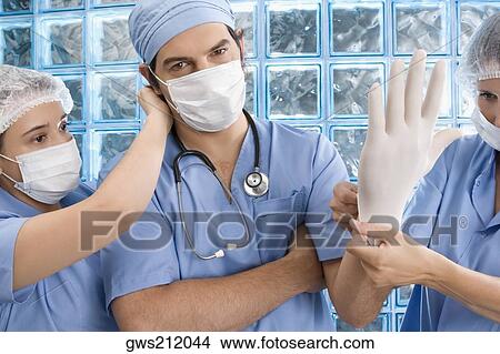 Picture - Two female surgeons adjusting surgical glove and surgical mask of a male doctor. Fotosearch