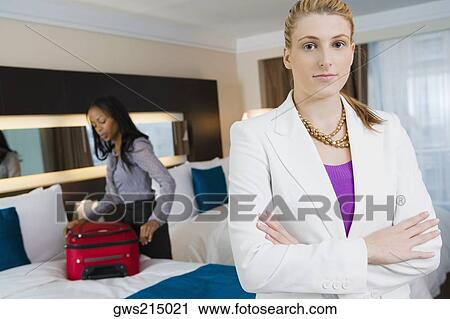 Stock Image - Portrait of a businesswoman standing with her arms crossed with another businesswoman packing her luggage. Fotosearch