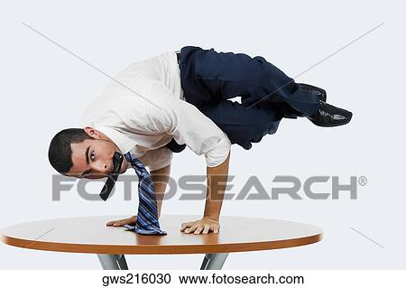 Stock Image - Portrait of a businessman doing a handstand on a table with a mobile phone in his mouth. Fotosearch