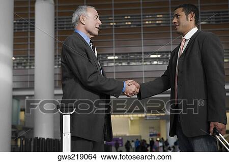 Picture - Side profile of two businessmen shaking hands at an airport lounge. Fotosearch