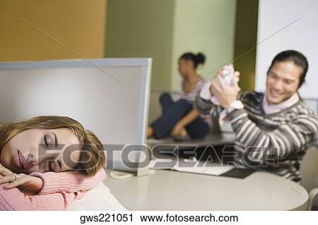 Stock Image - Young woman napping on a desk with a young man smiling in the background. Fotosearch