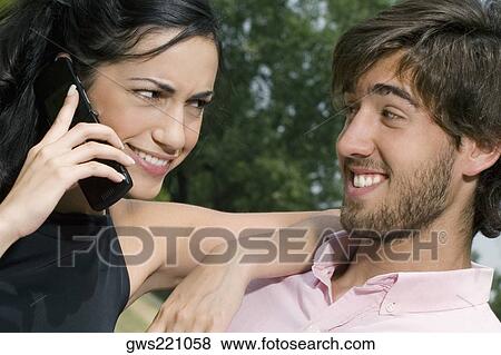 Young woman talking on a mobile phone with a young man smiling View Large Photo Image Stock Photo - Young woman talking on a mobile phone with a young man smiling. Fotosearch