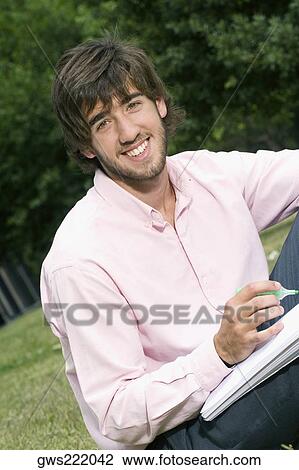 Stock Image - Portrait of a young man writing with a pen on a notepad and smiling. Fotosearch