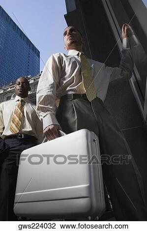 Stock Image - Low angle view of a businessman holding a briefcase and another businessman standing behind him. Fotosearch